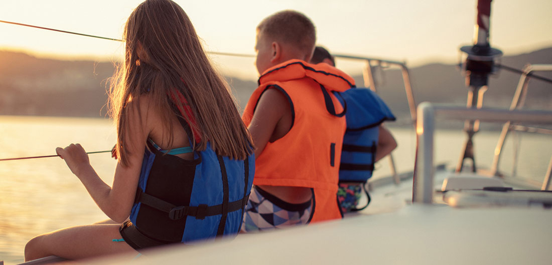 two children, girl and boy, wear life jackets on deck of boat looking at sunset