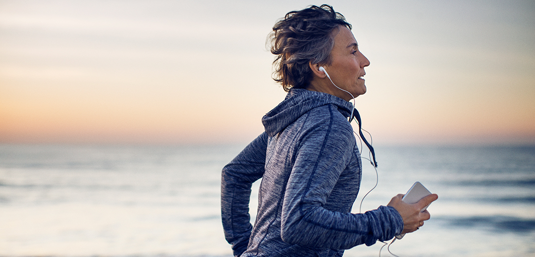 middle aged woman listens to headphones while she runs on the beach at sunset