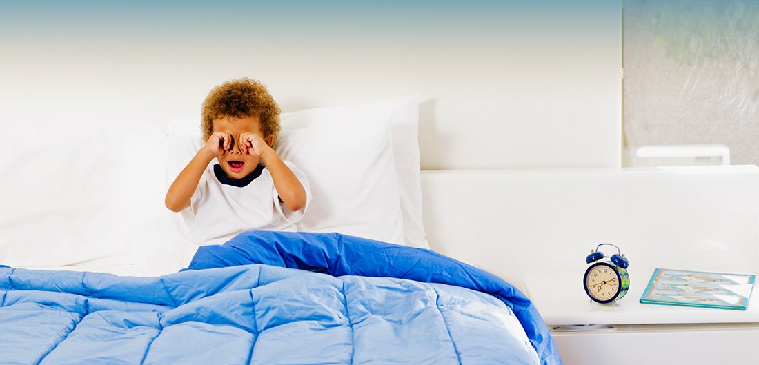 little boy rubbing his eyes in bed with a clock next to him