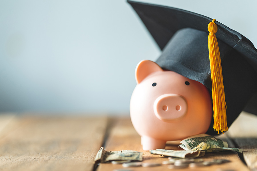 Apply For The 2023 Miller’s Insurance Agency Scholarship - Piggybank Wearing a Graduation Cap Sitting on a Wooden Desk Next to Money
